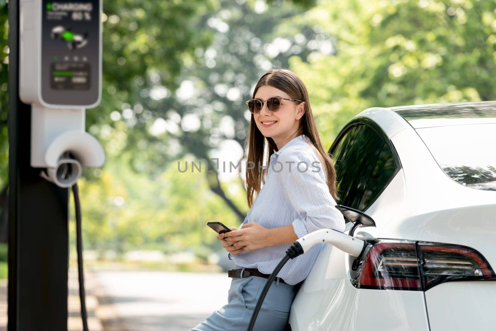 Young woman using smartphone online banking application to pay for electric car battery charging from EV charging station during vacation holiday road trip at national park or summer forest. Exalt