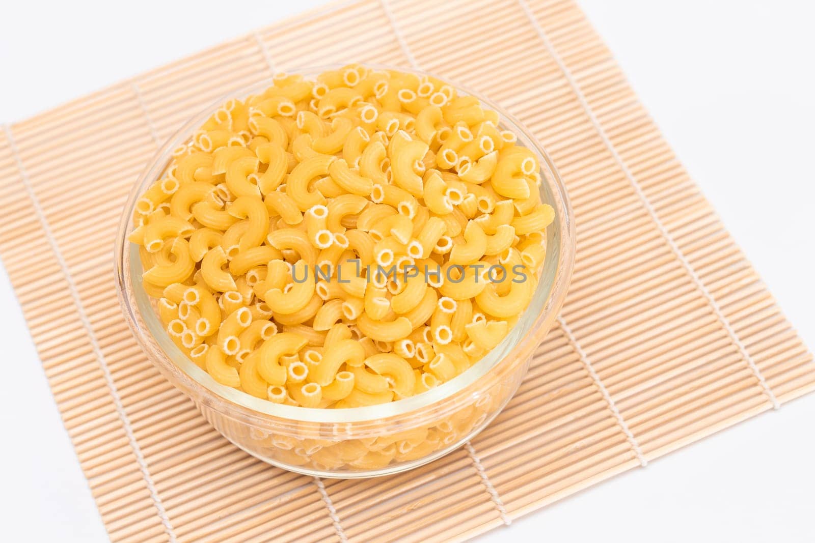 Uncooked Chifferi Rigati Pasta in Glass Jar on Bamboo Mat on White Background. Fat and Unhealthy Food. Classic Dry Macaroni. Italian Culture and Cuisine. Raw Pasta