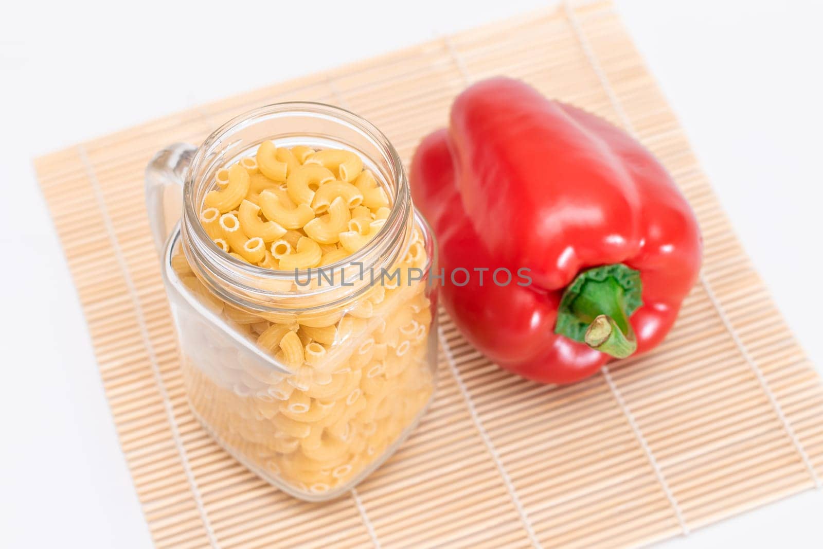 Uncooked Chifferi Rigati Pasta in Glass Jar and Red Bell Pepper on Bamboo Mat on White Background. Fat and Unhealthy Food. Classic Dry Macaroni. Italian Culture and Cuisine. Raw Pasta