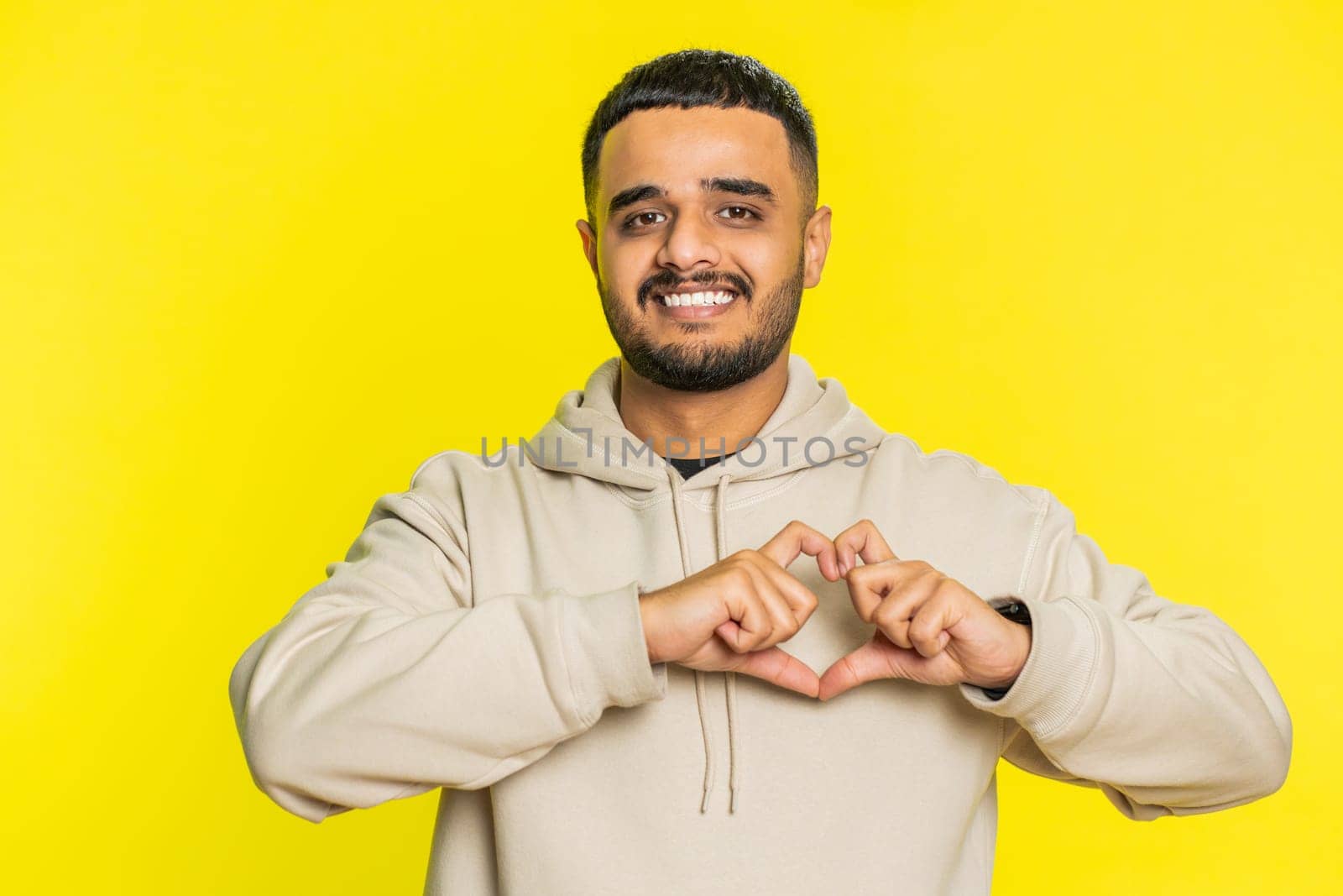 Man in love. Smiling attractive Indian man makes heart gesture demonstrates love sign expresses good positive feelings and sympathy. Handsome Arabian Hindu young guy isolated on yellow wall background
