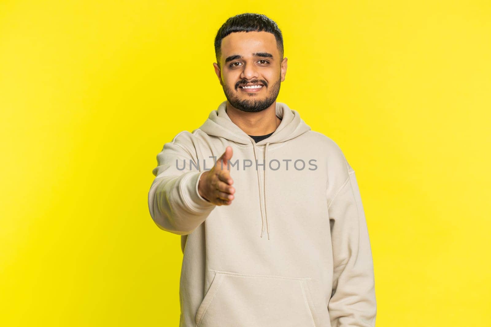 Friendly Indian young man outstretching hand to camera, offering handshake, greeting with kind smile, invitation, welcoming showing solidarity, trust concept. Arabian guy isolated on yellow background