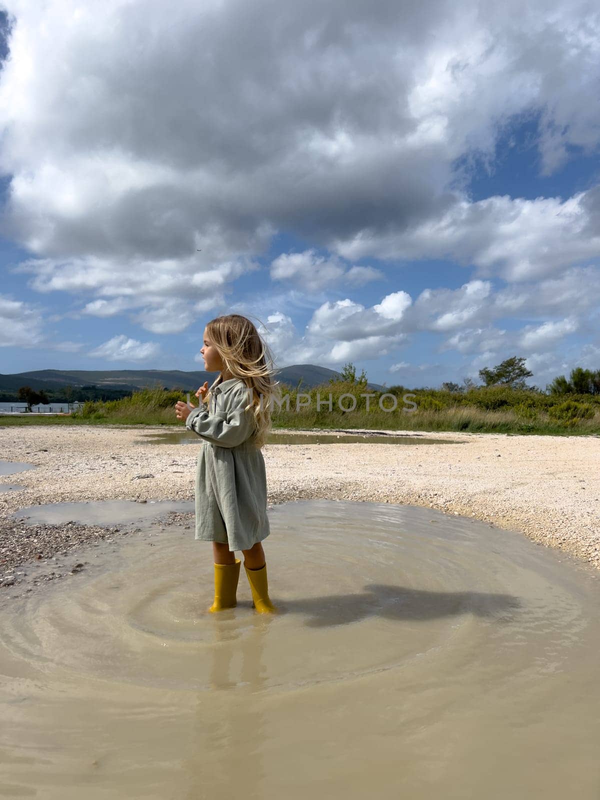 Little girl with flying hair stands sideways in a puddle in rubber boots. High quality photo