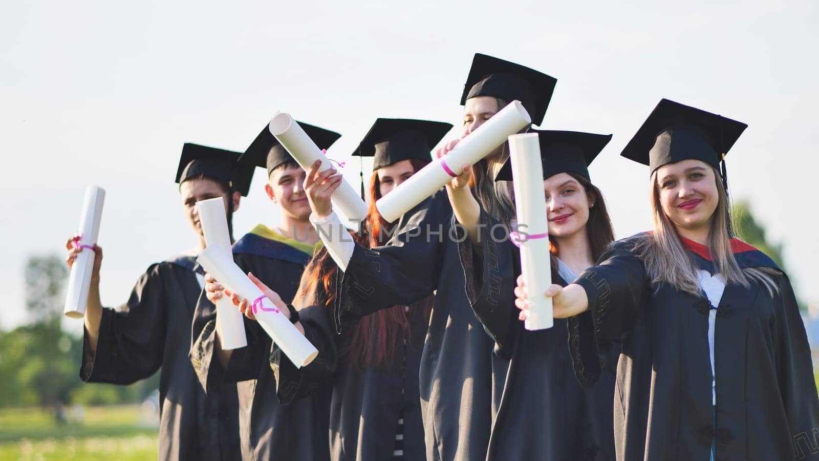 Cheerful graduates pose with raised diplomas on a sunny day. by DovidPro