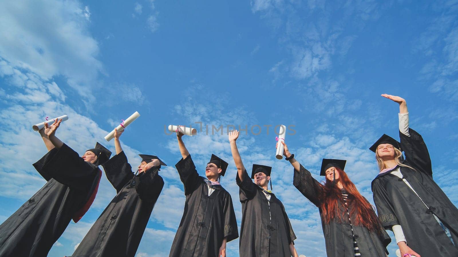 Cheerful graduates pose with raised diplomas on a sunny day