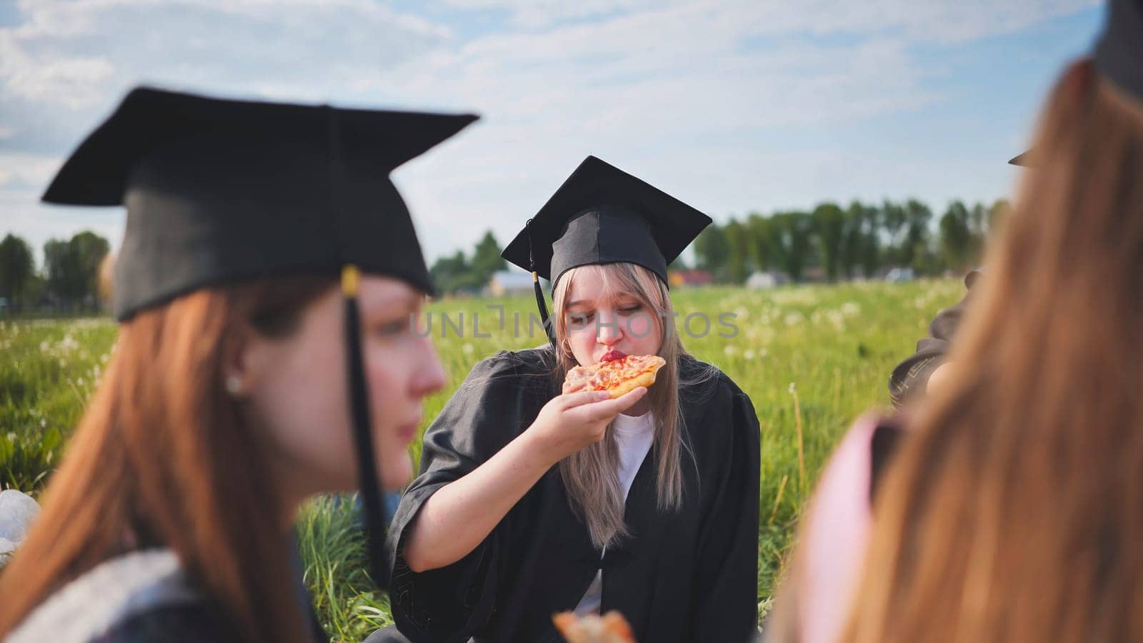 Graduates in black suits eating pizza in a city meadow