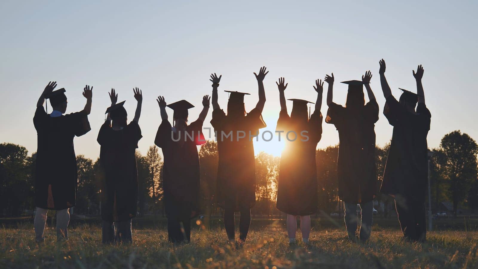 College graduates in robes waving at sunset. by DovidPro