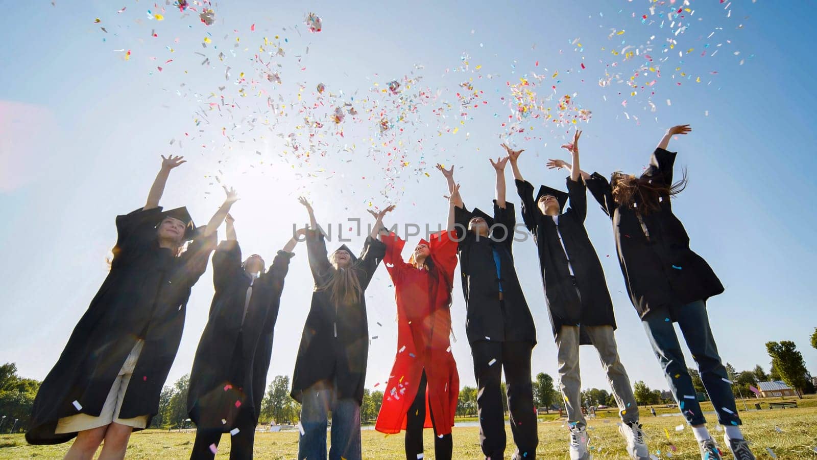 Happy college graduates throw colorful confetti against the rays of sunshine. by DovidPro