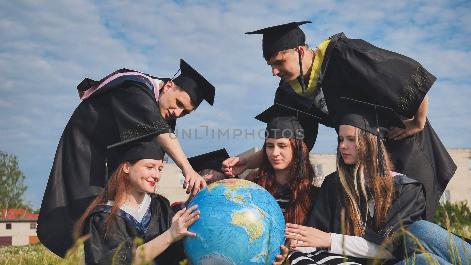 Graduates in black robes examine a geographical globe sitting on the grass. by DovidPro