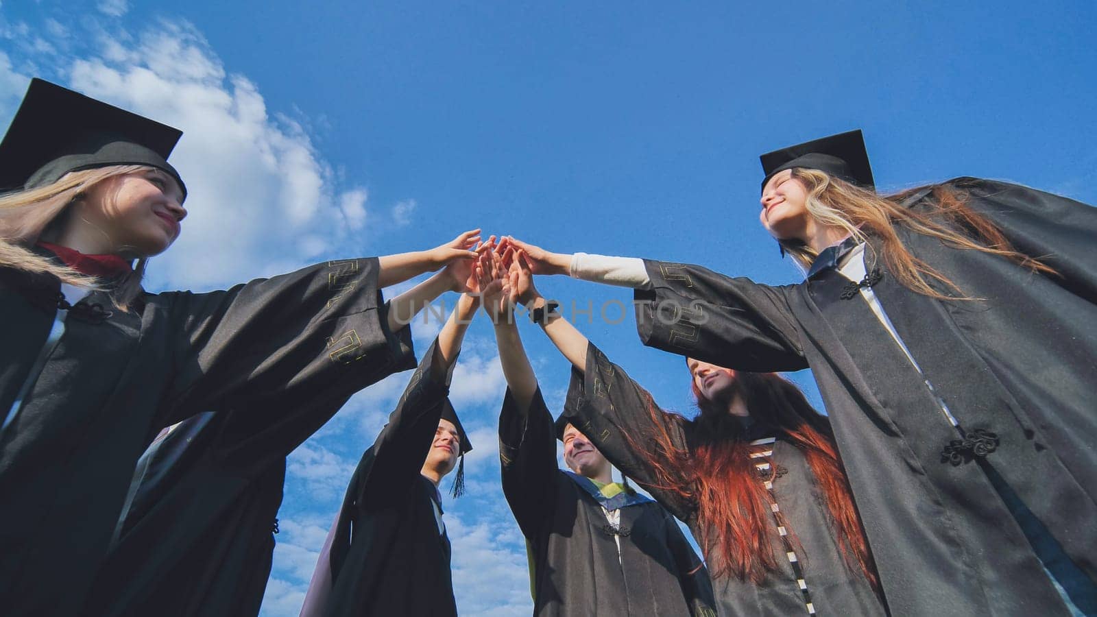 Group of happy successful graduates in academic hats and robes standing in circle and putting their hands together. Team of college or university students celebrating graduation. by DovidPro