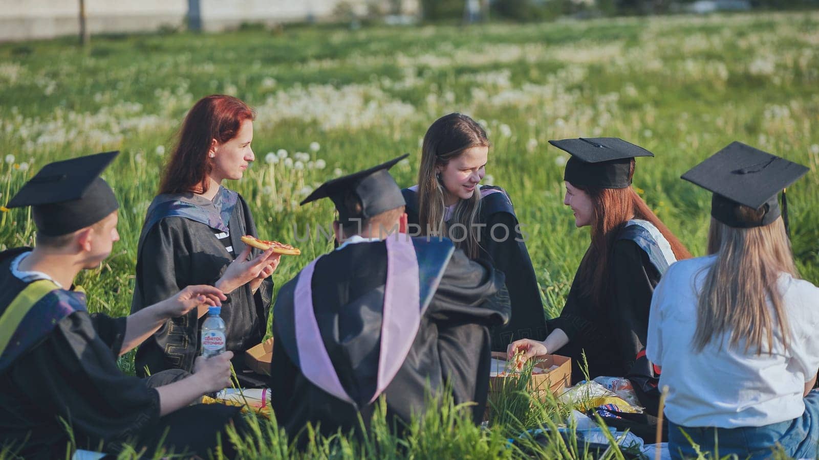 Graduates in black suits eating pizza in a city meadow