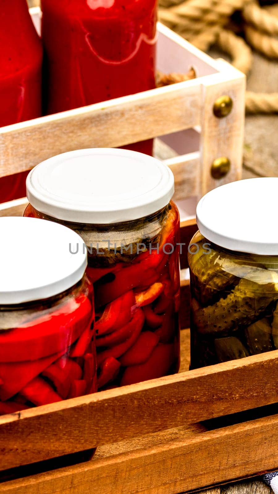 Wooden crate with glass jars with pickled red bell peppers and pickled cucumbers (pickles) isolated. Jars with variety of pickled vegetables. Preserved food concept in a rustic composition.