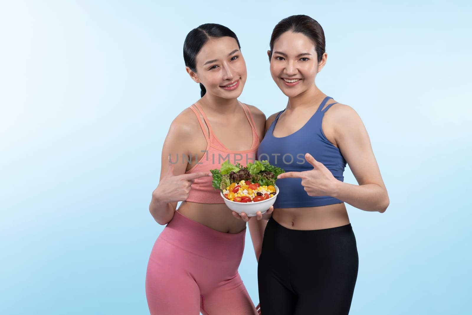 Two young sporty Asian women in sportswear holding salad bowl fill with fruit and vegetable. Natural youthful and fit body lifestyle people with balance nutrition on isolated background. Vigorous