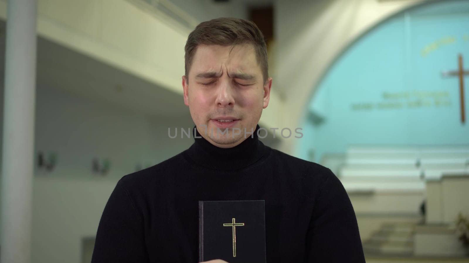 A young man prays holding a bible in his hands in a church against the background of a cross. A Protestant man stands in a church and prays. 4k