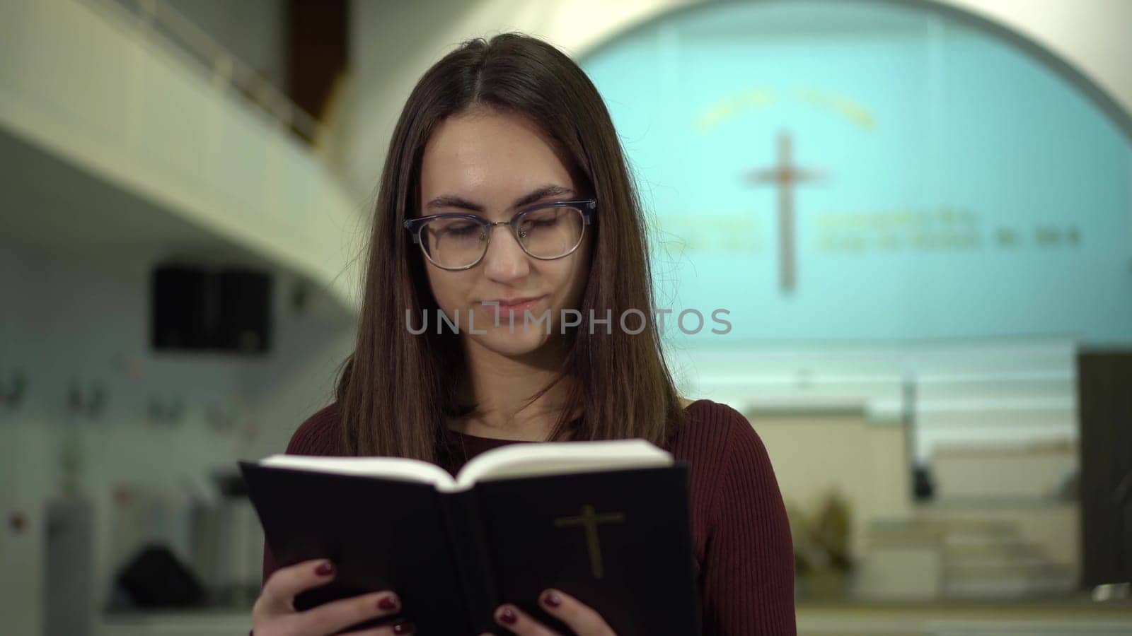A young woman reads the Bible against the background of a cross in a church. A Protestant girl with glasses reads the Bible in church closeup. by Puzankov