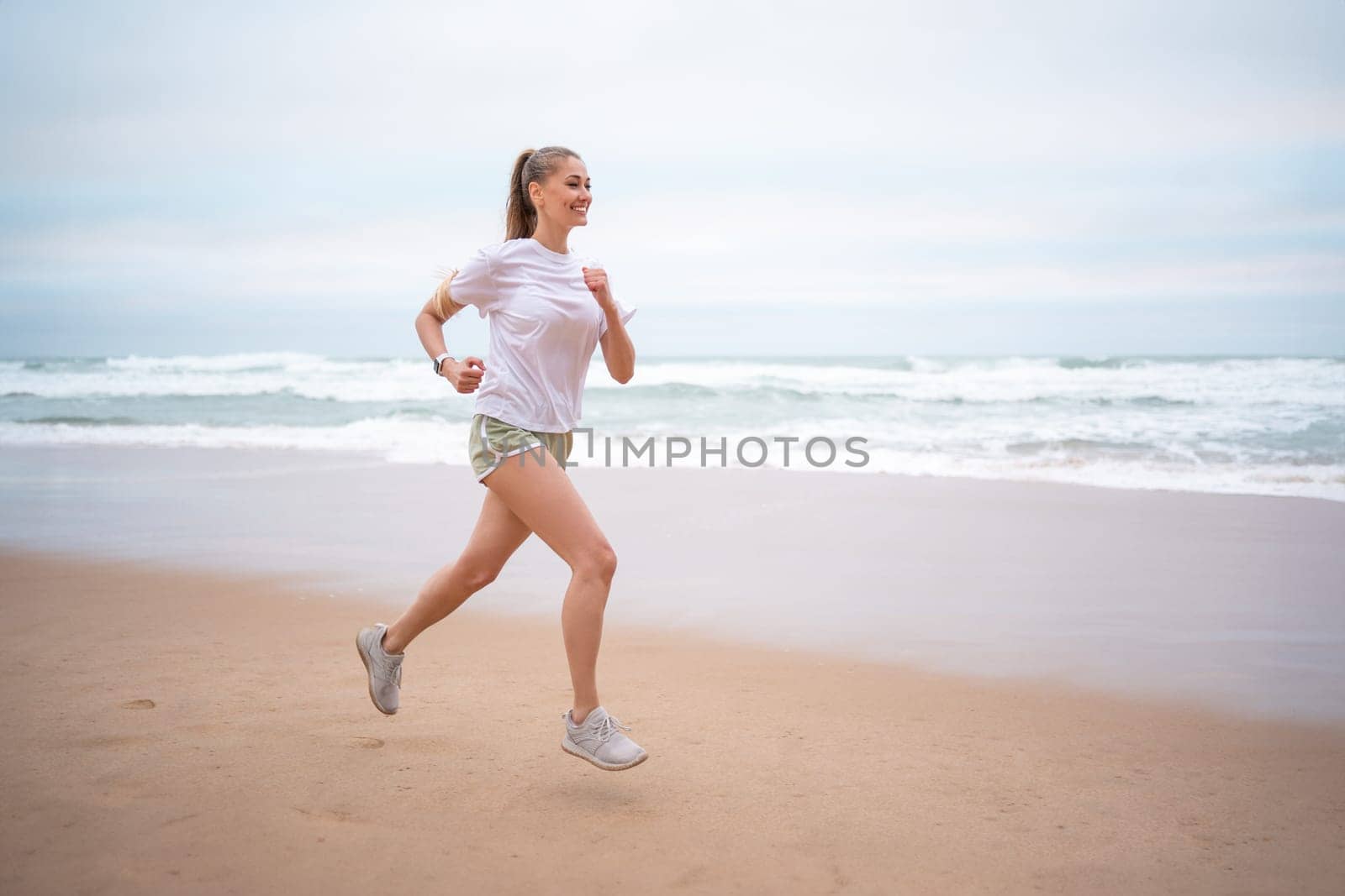 Woman running along ocean shore beach. Jogging on nature Woman in white shirt top and green sports shorts runs along seashore. Athletic woman run along sandy beach on seashore in morning