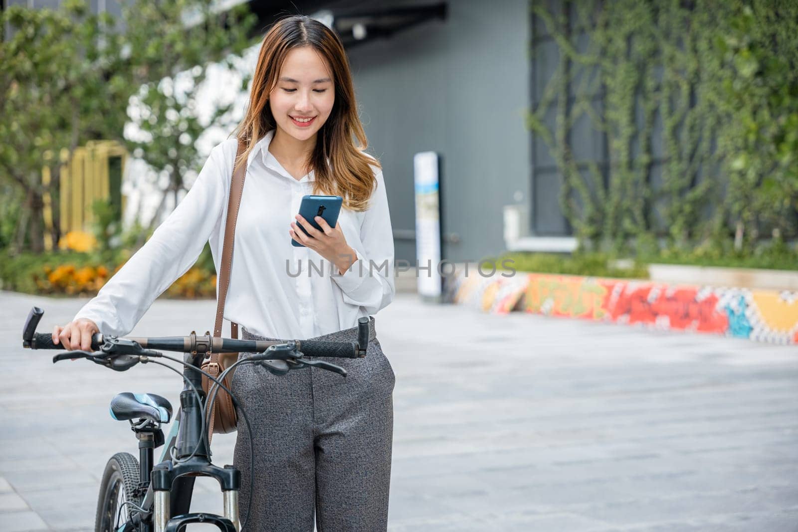 businesswoman standing on city street building with bicycle holding smart mobile phone by Sorapop