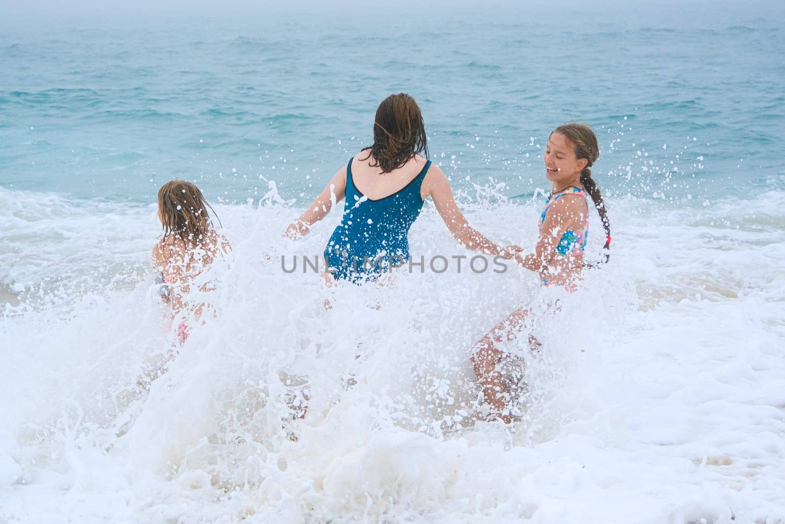 Mother and children playing on the ocean beach. Family enjoying the ocean. Mother holds girls's hands and they all look at the ocean together.