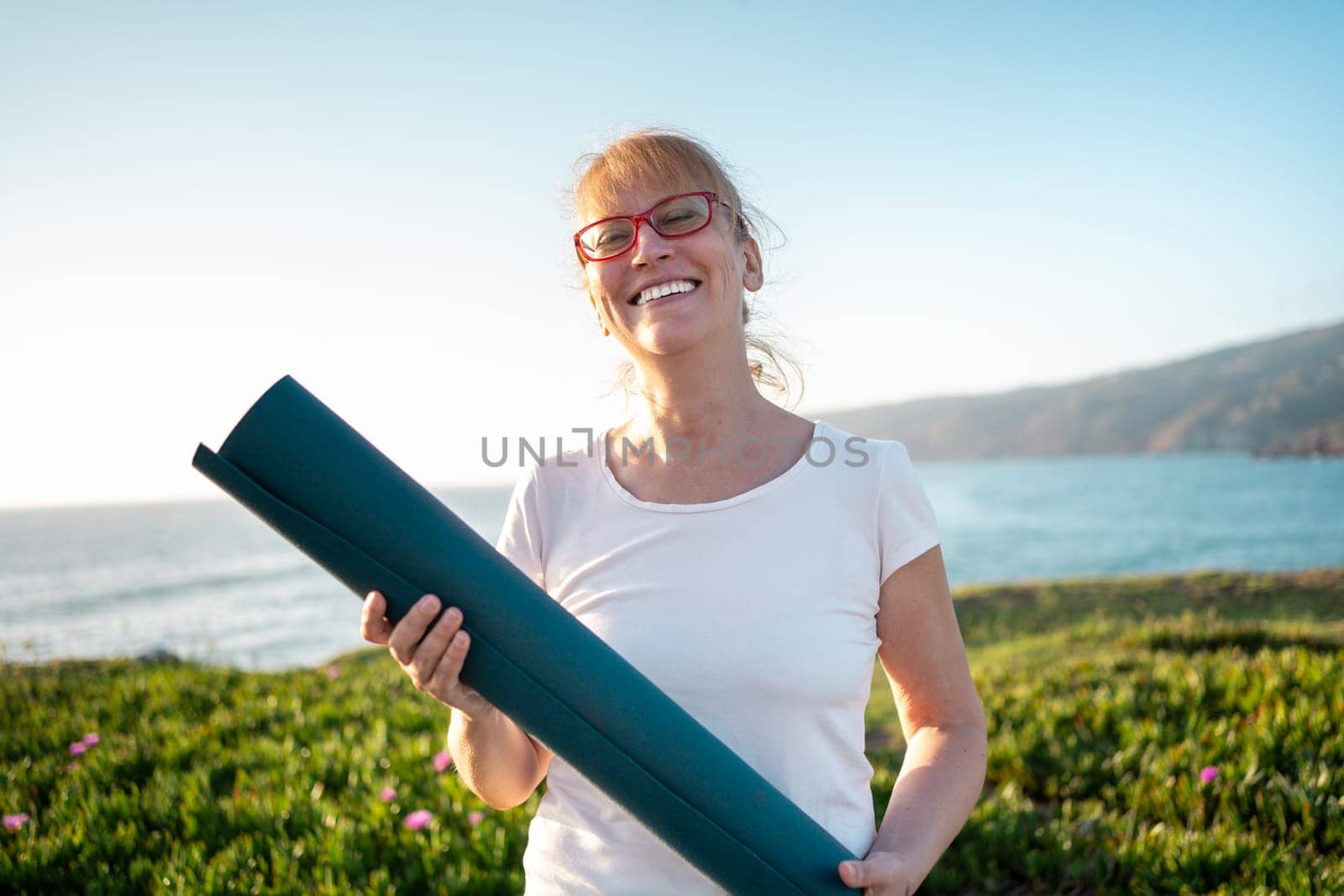 Happy senior woman wear glasses holding yoga mat smiling on ocean shore by andreonegin