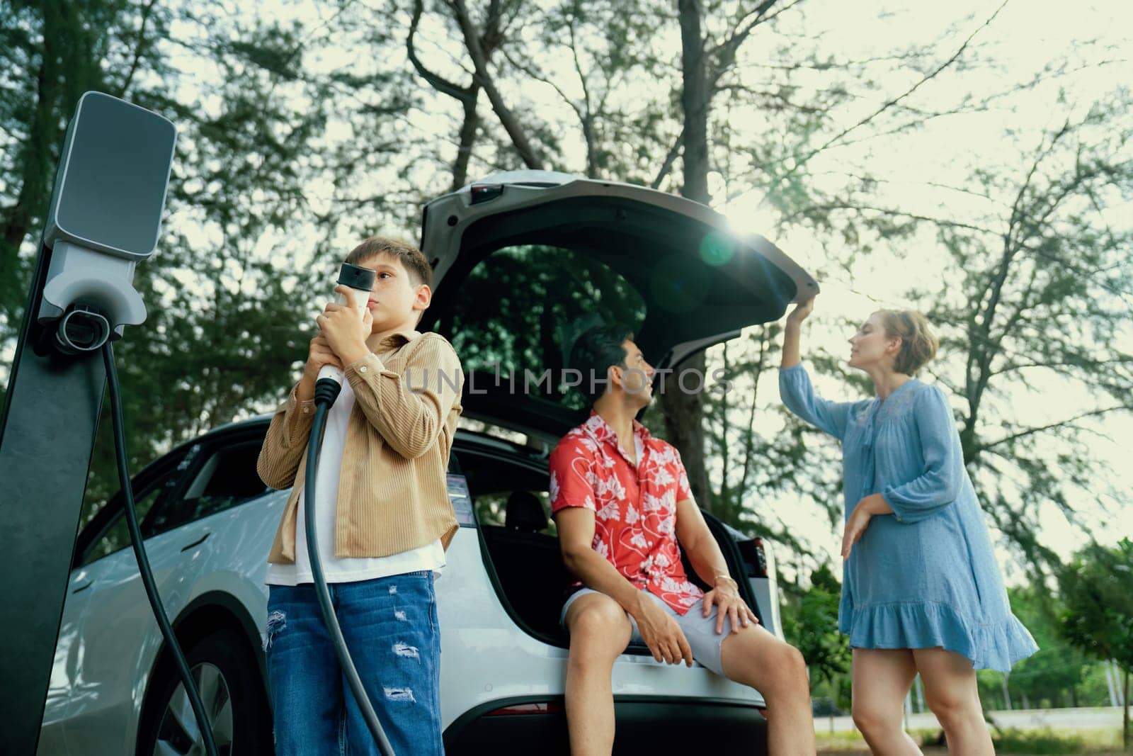 Little boy holding EV charger and point at camera with his family sitting on the trunk in background. Road trip travel with alternative energy charging station for eco-friendly car concept. Perpetual