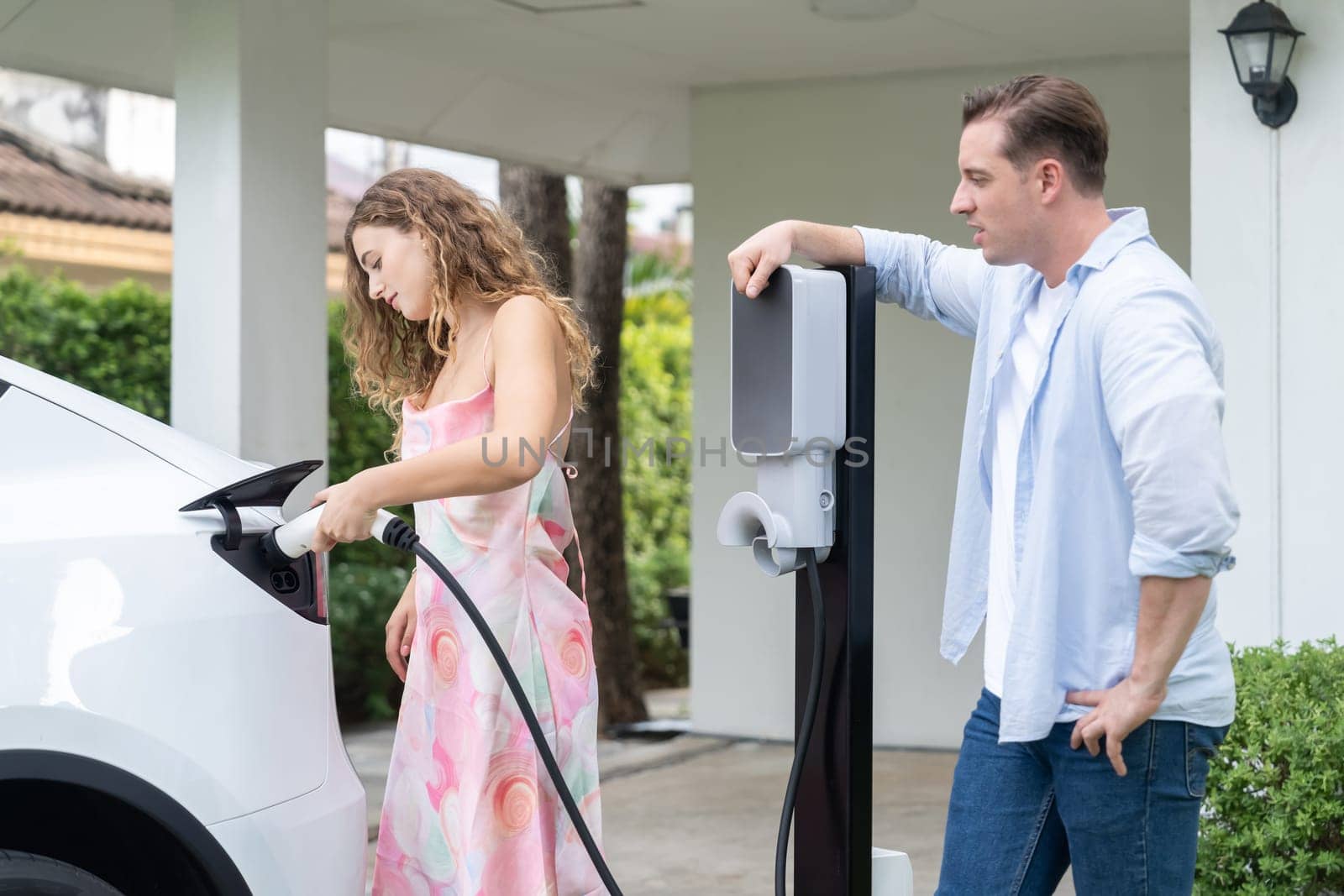 Happy and lovely couple with eco-friendly conscious recharging electric vehicle from EV home charging station. EV car technology utilized for residential home to future sustainability. Synchronos