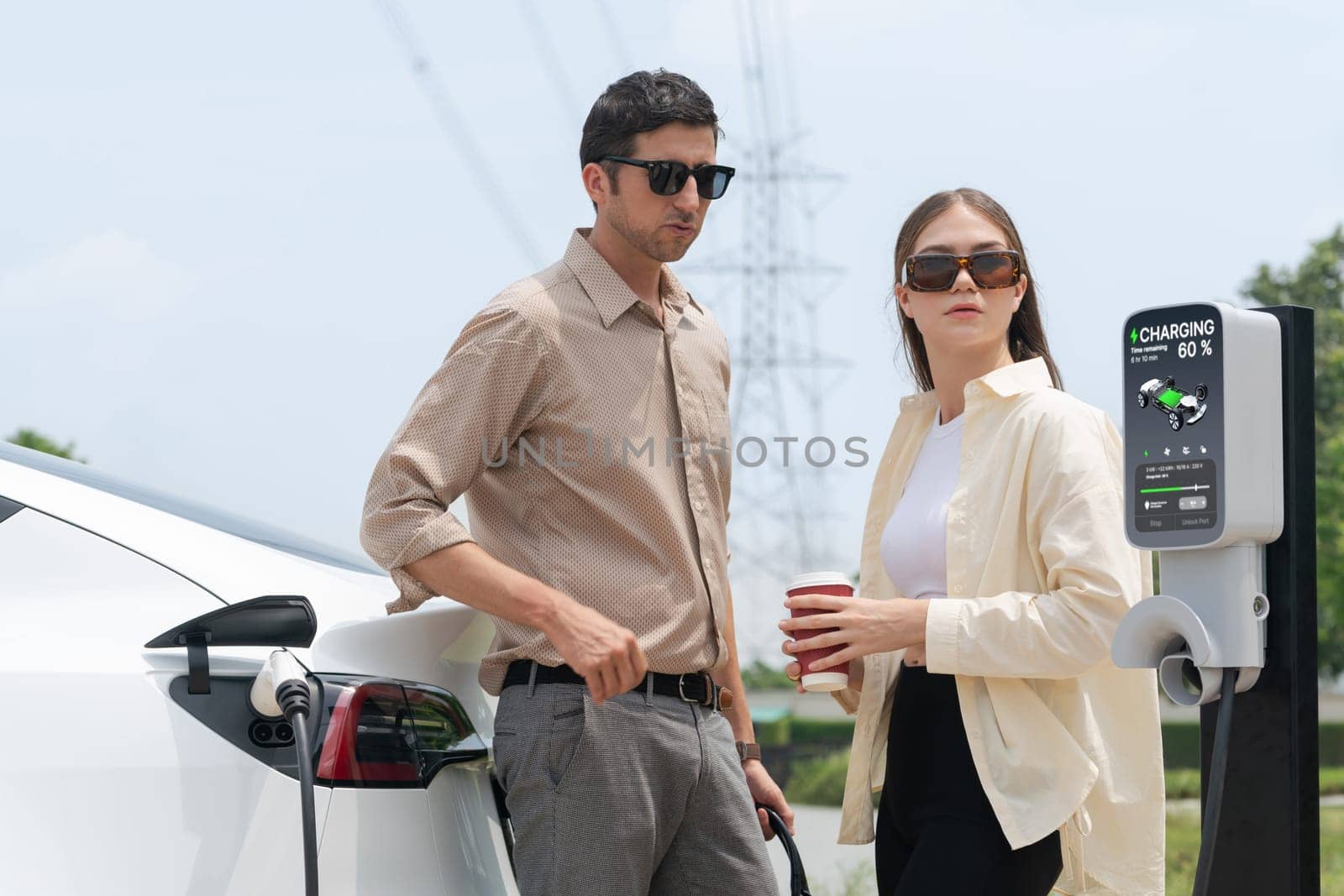 Young couple with coffee recharge EV car battery at charging station connected to electrical industrial power grid tower. Couple with shopping and travel using eco electric car lifestyle. Expedient
