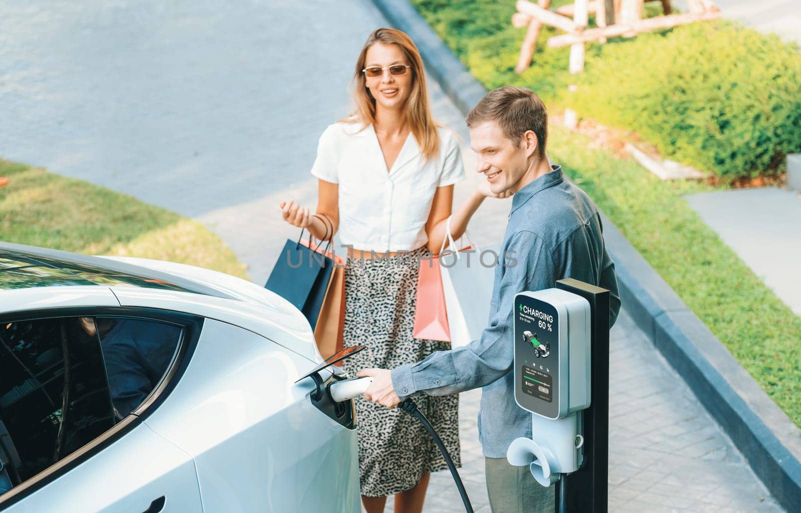 Young couple travel with EV electric car charging in green sustainable city outdoor garden in summer shows urban sustainability lifestyle by green clean rechargeable energy of electric vehicle innards