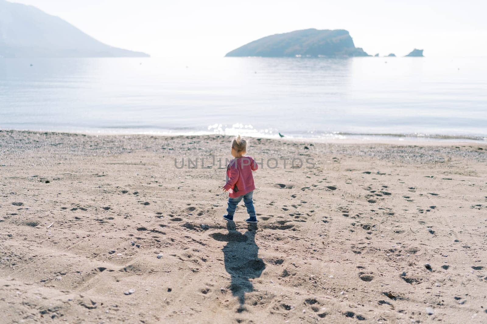 Little girl walks along the sandy beach near the sea. Back view by Nadtochiy