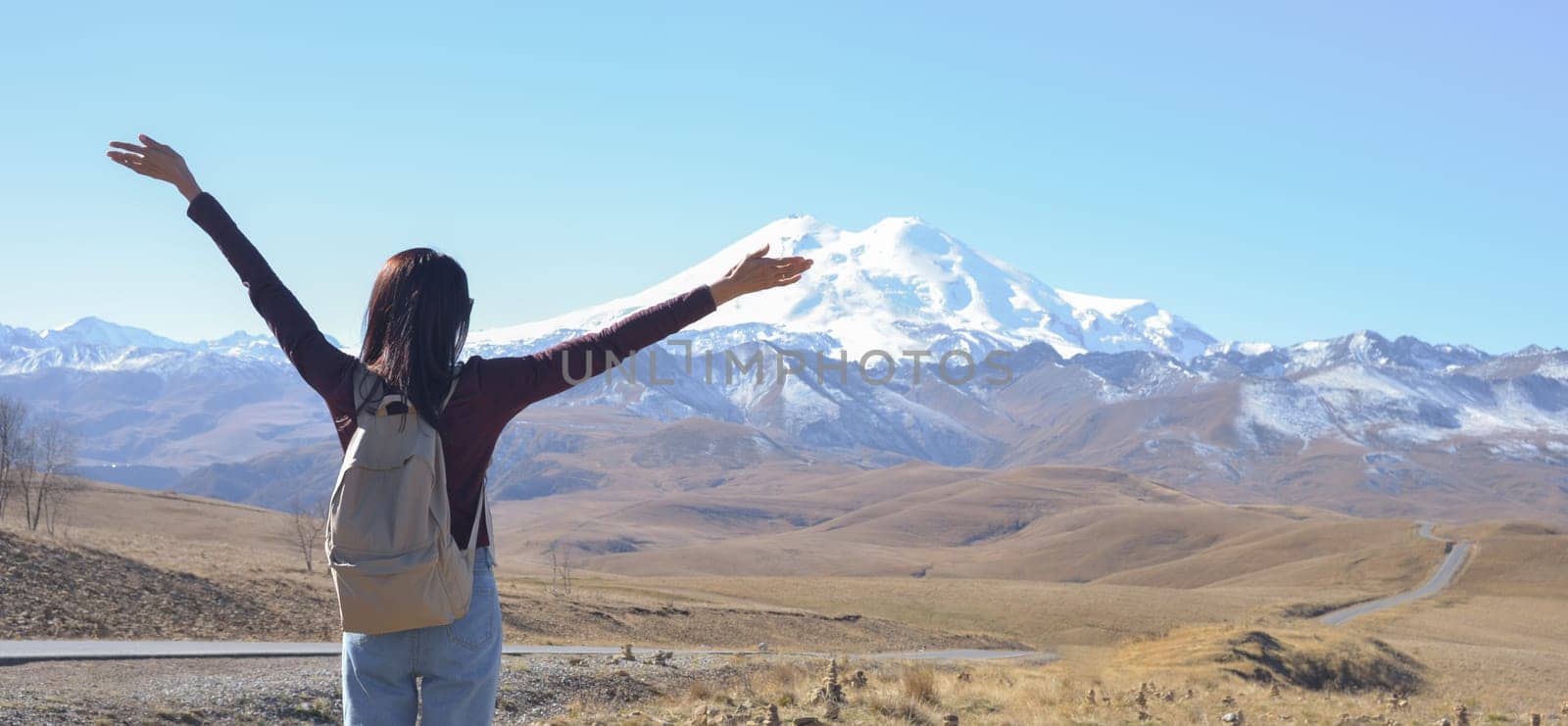 A happy female traveler with a backpack behind her back and her arms raised up admires the snow-covered Elbrus on a clear autumn day. View of Elbrus, North Caucasus, Russia by Ekaterina34