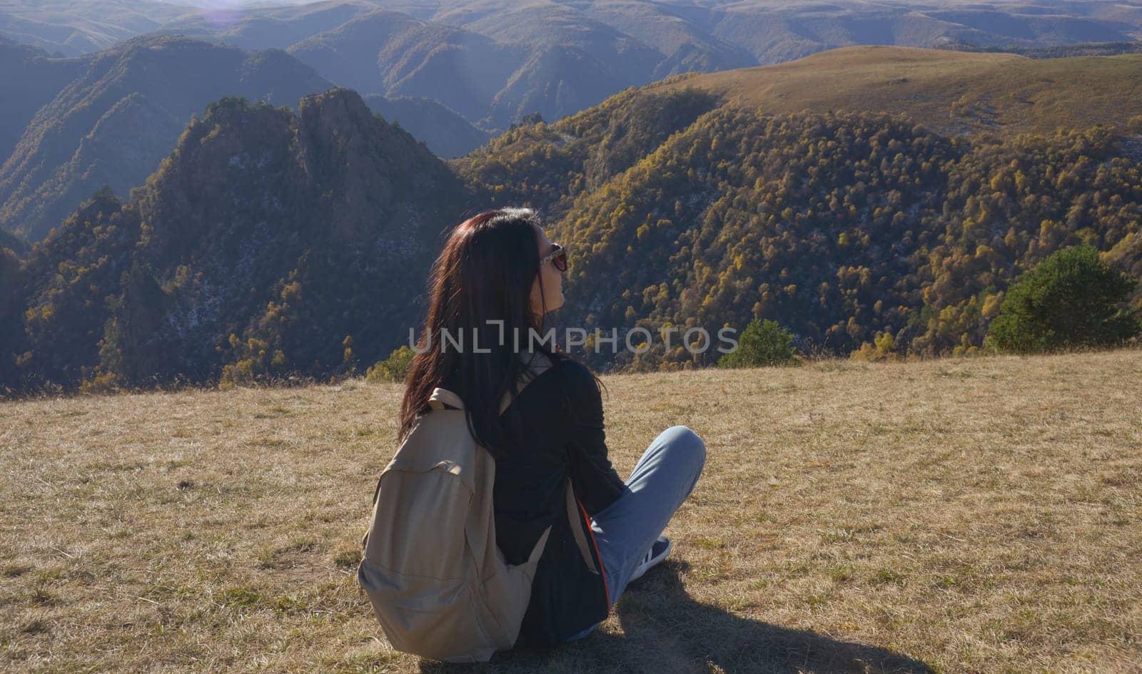 A female traveler with a backpack behind her sits on the grass and enjoys the views of Elbrus by Ekaterina34