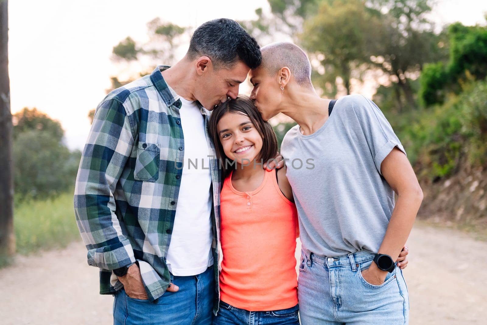 portrait in the countryside of a father and mother kissing their daughter who smiles happily looking at camera, concept of active tourism in nature and family outdoors activities