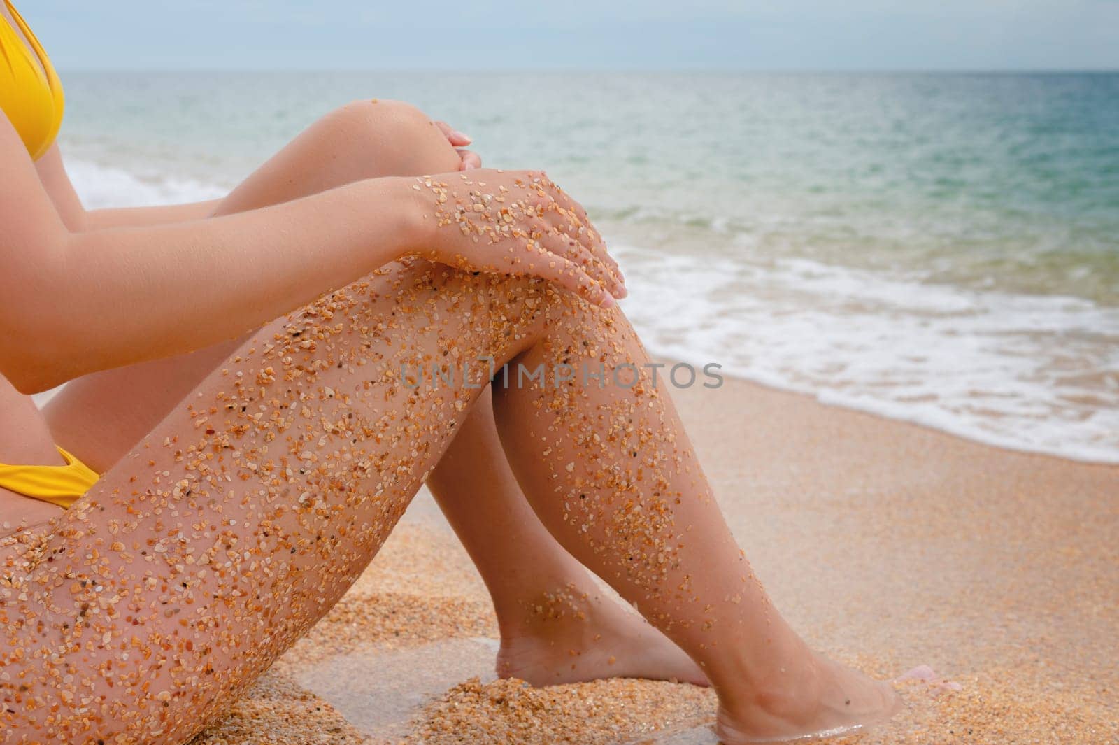 Close-up of sea waves near the feet of a young woman who slowly sits and relaxes on the beach. Women's feet on the sea sand.