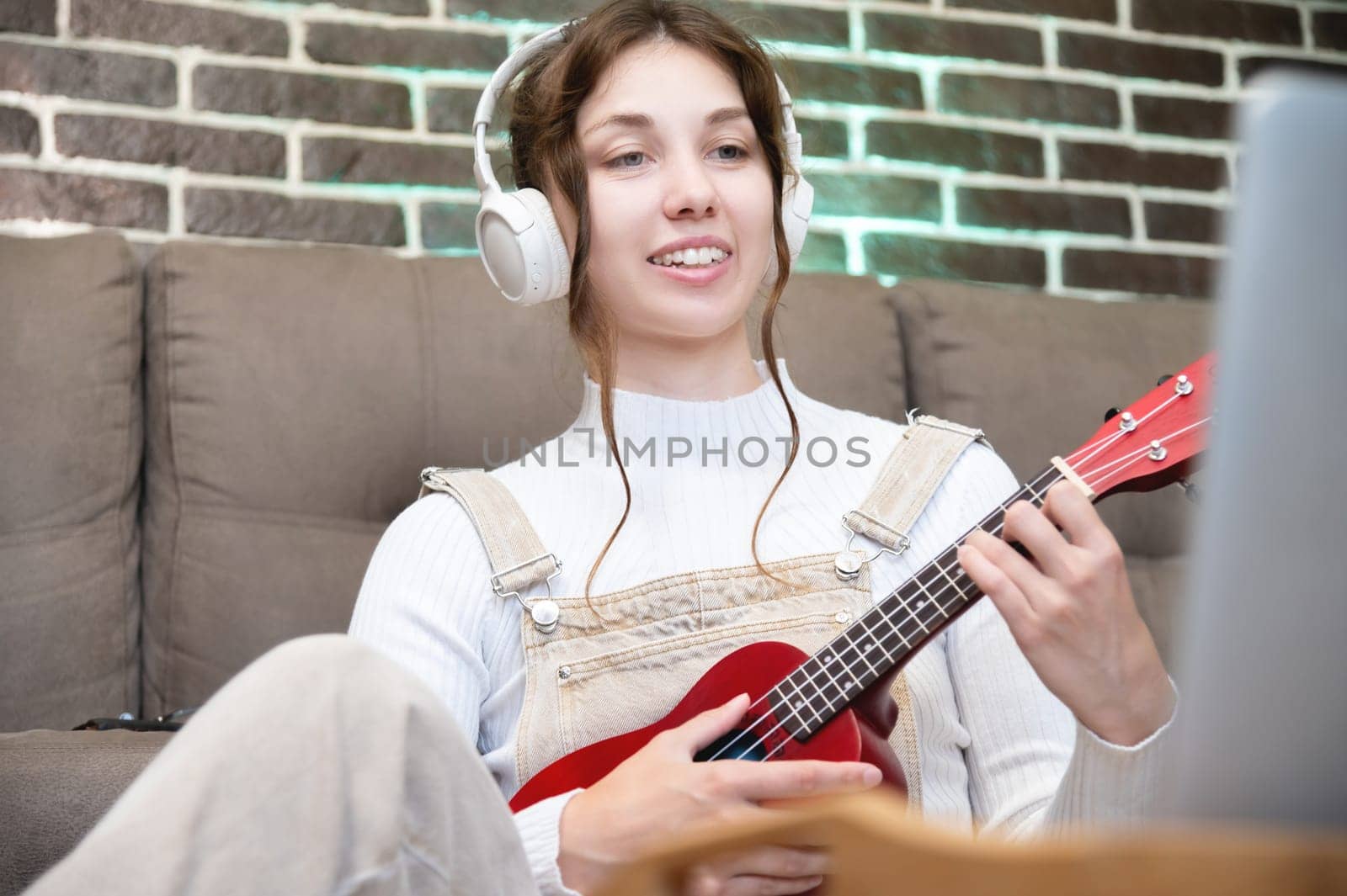 young cheerful girl in a sweater is learning to play the ukulele. Online learning, home education concept. Learning new skills by yanik88