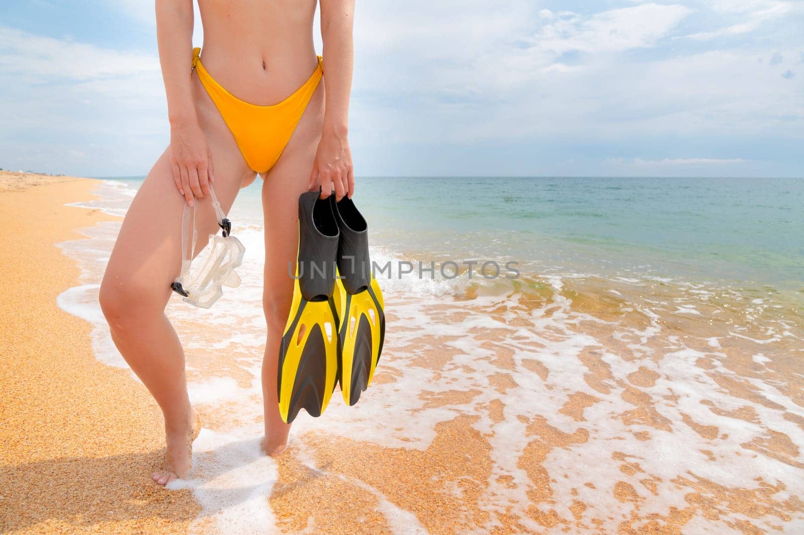 relaxed girl stands with snorkeling equipment in her hands on the seashore. view of the legs of a young woman in a swimsuit by yanik88