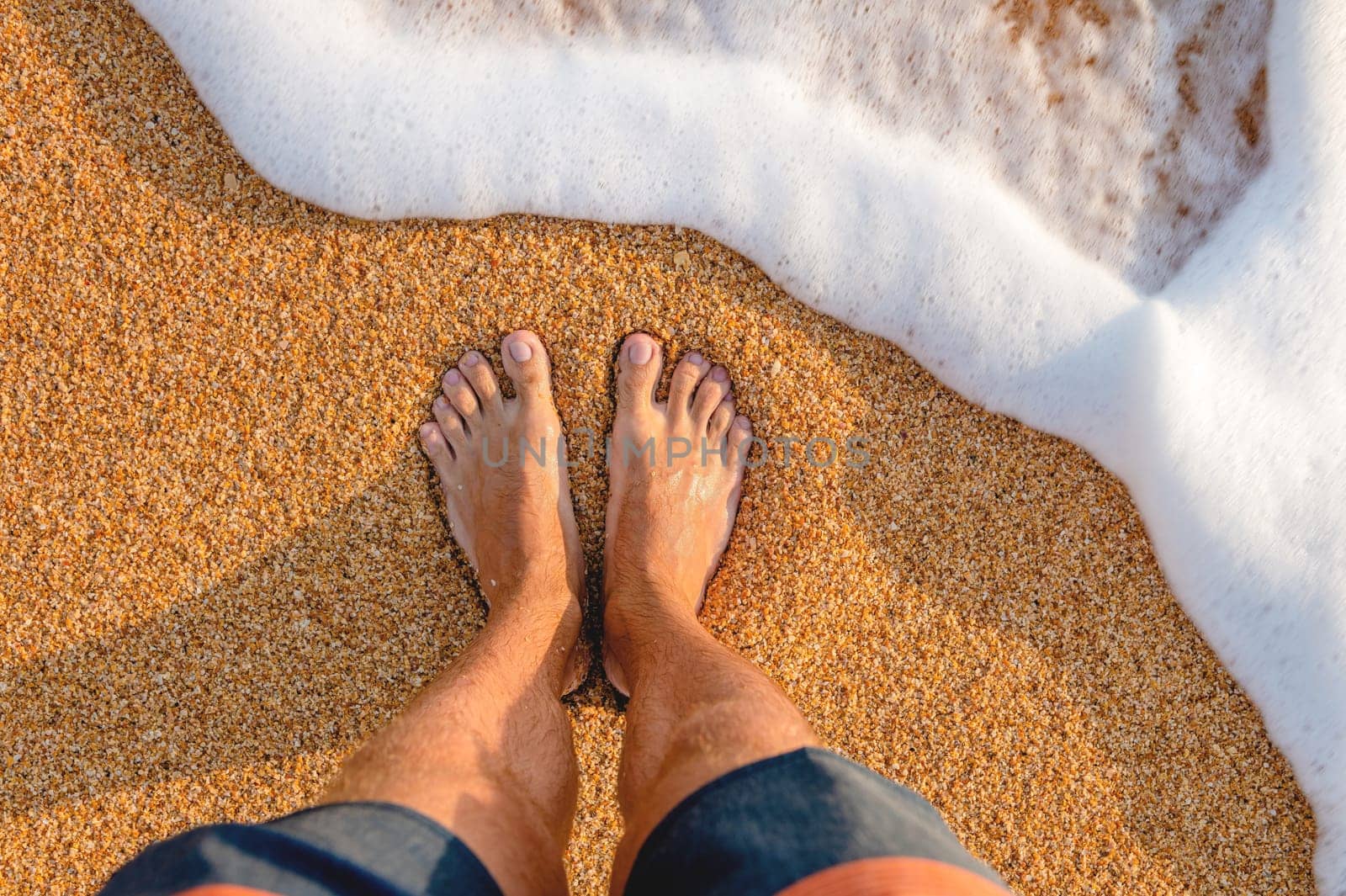 male feet barefoot on the waves of sea foam on a sandy golden beach on a summer day. top view of male legs in shorts and clear waters of the ocean by yanik88