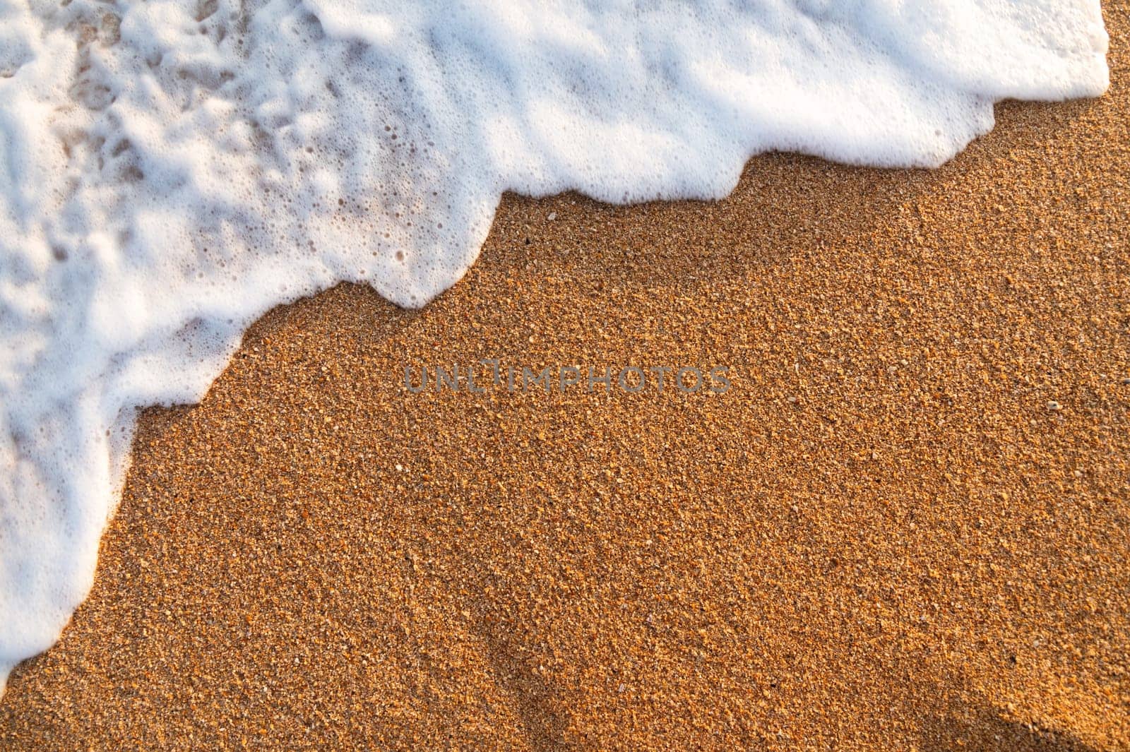 a wave rolls onto the sea beach with a splash. close-up white abundant foam lies on golden sandy sand, top view