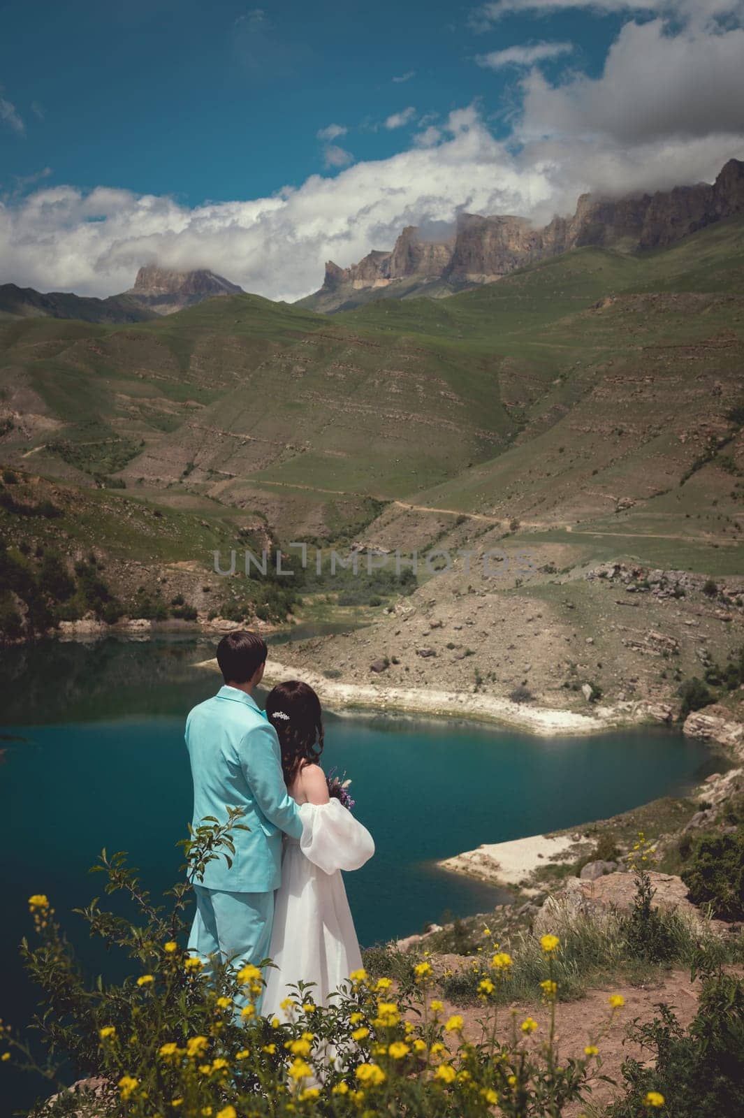 Bride and groom dressed elegant in their wedding gowns holding hands and hugging posing for their photo session in a desert like landscape by the lake by yanik88