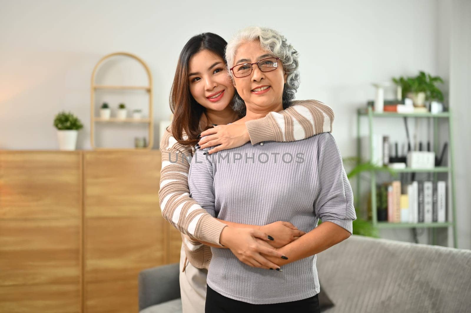 Young Asian woman hugging happy mature mother from back while standing in living room. Family bonding concept.