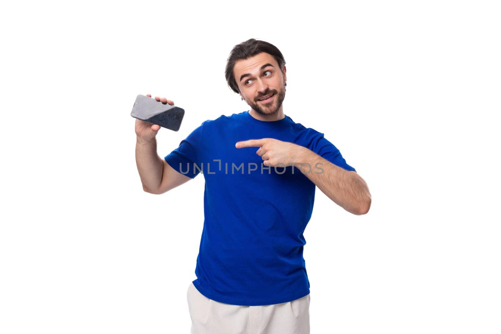 young attractive brunette man with a beard in a blue t-shirt holding a smartphone with a mockup on a white background with copy space.