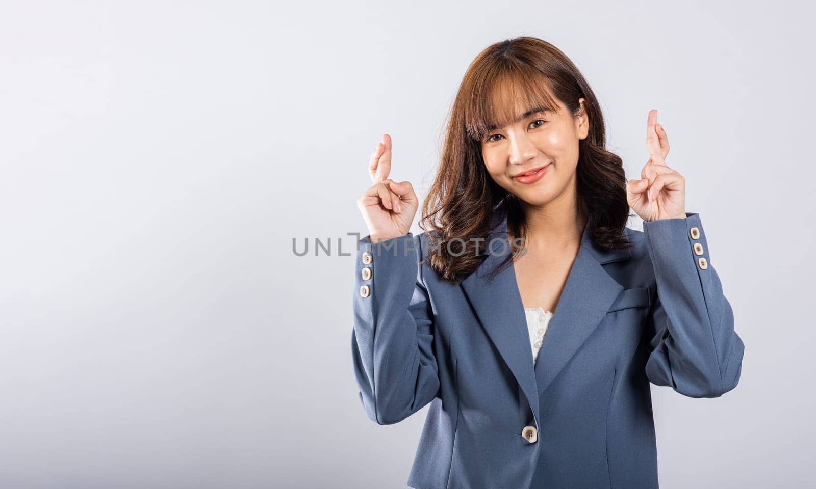 Superstition meets joy in this studio shot of a cute Asian lady. She holds crossed fingers, symbolizing hope and positivity. Isolated on a white background, her expressive face shines with happiness.