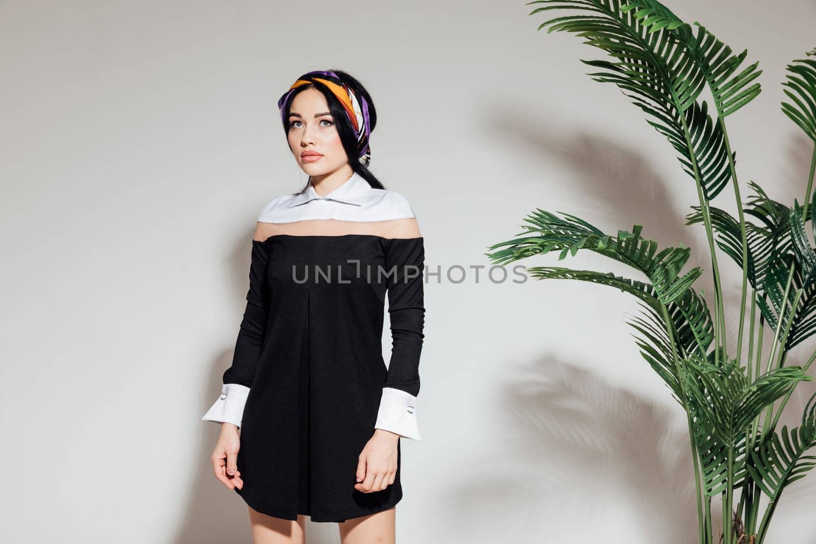 a woman in a black dress and scarf stands against a white wall and a green plant