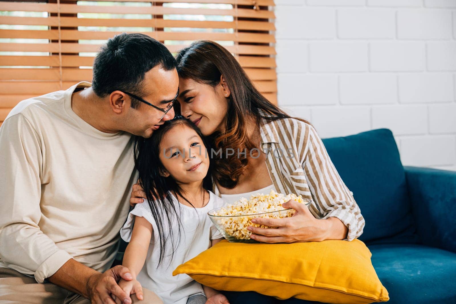 A happy family filled with joy watches TV while eating popcorn on modern sofa at home by Sorapop