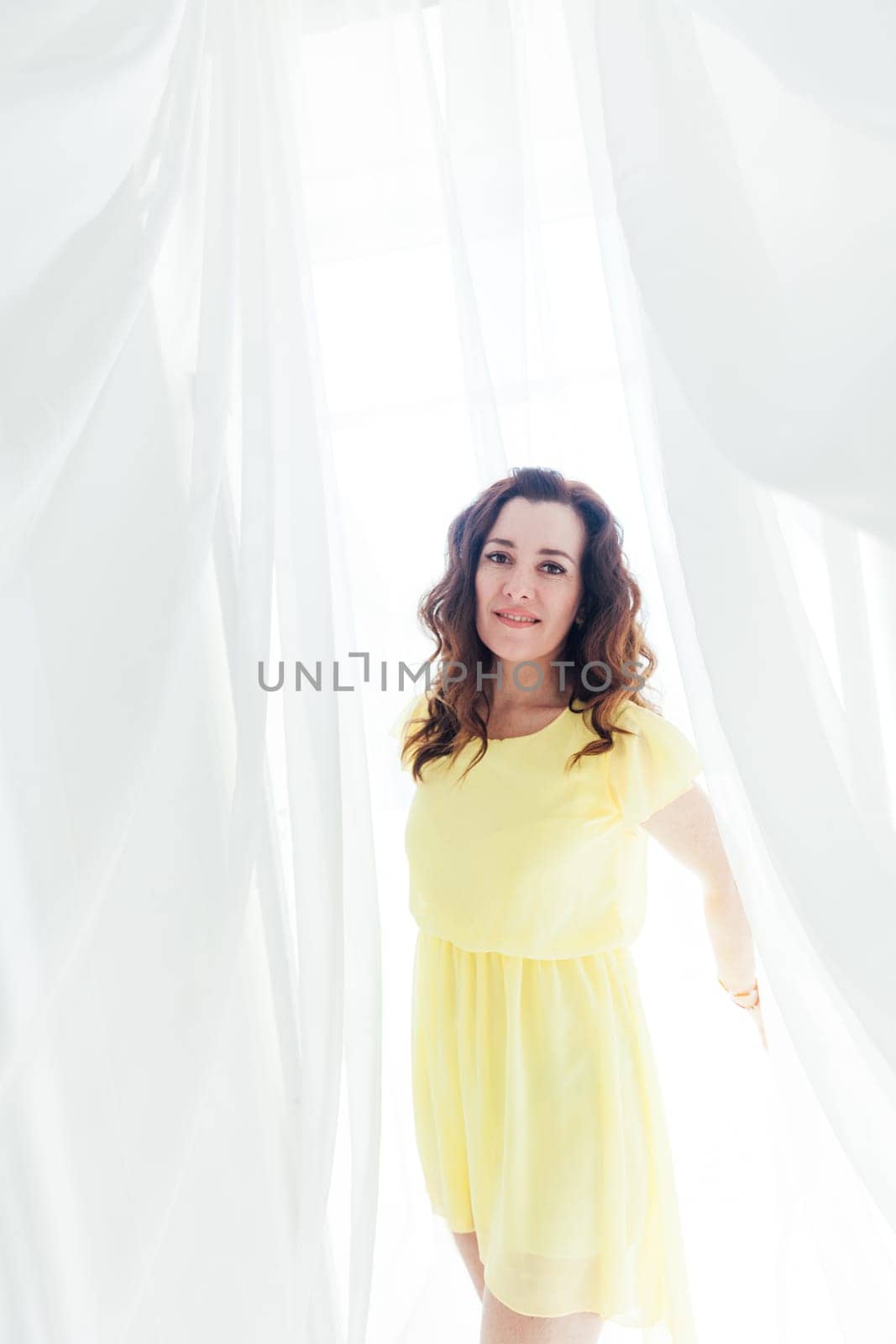 beautiful brunette woman in yellow dress stands at the white curtains of tulle fabrics in a bright room
