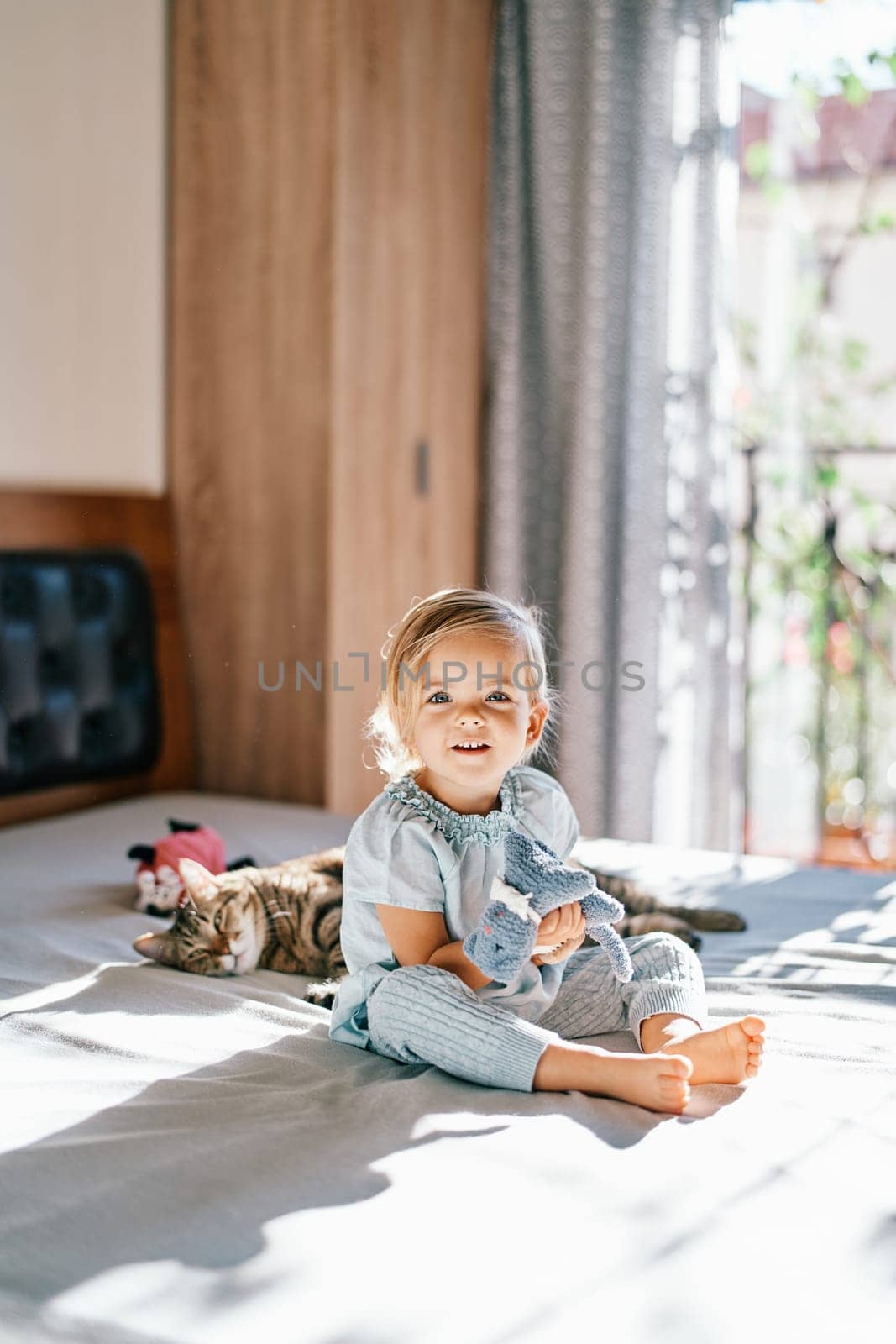 Little smiling girl with a soft toy in her hand sits on the bed next to a sleeping tabby cat by Nadtochiy