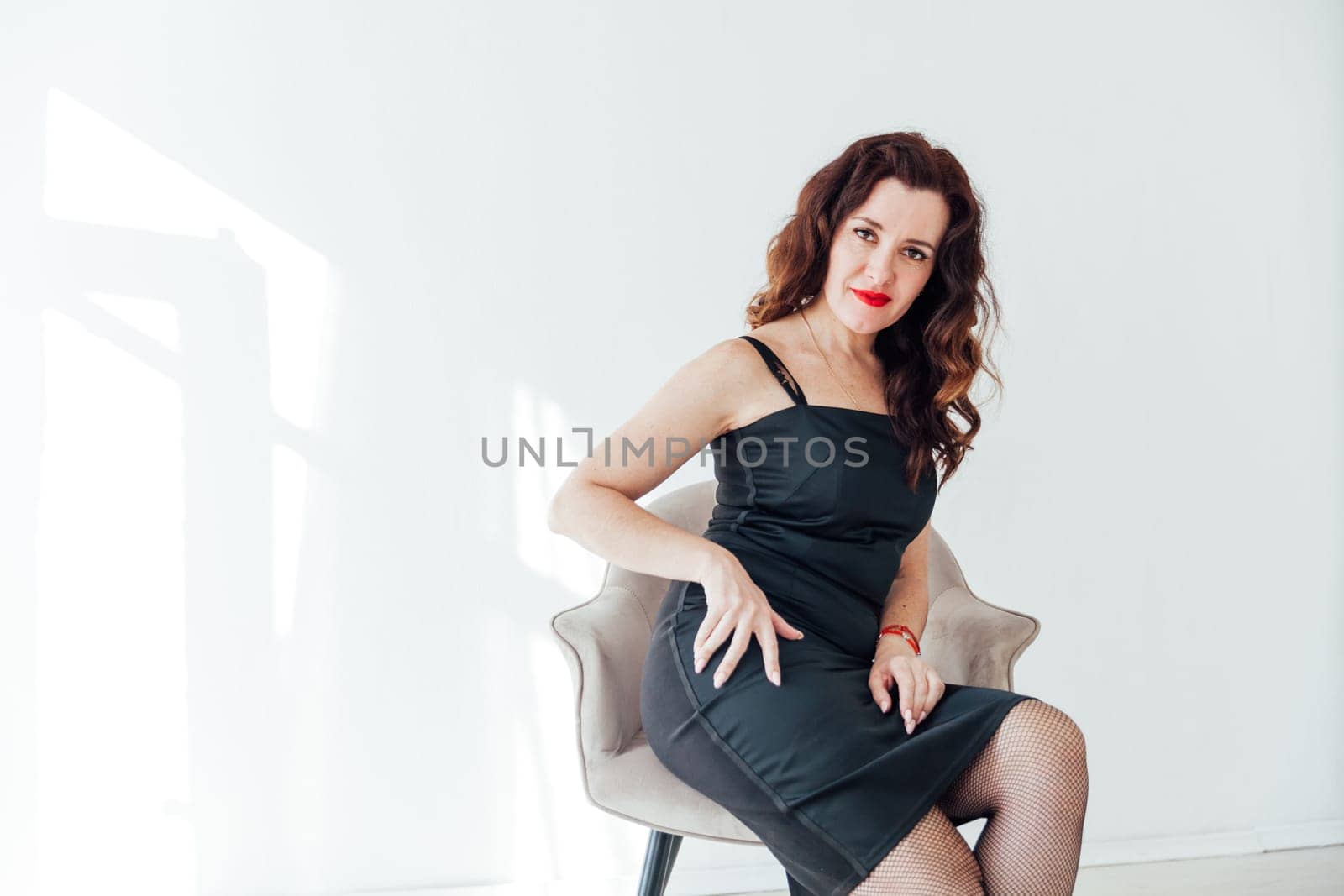 a brunette in a black dress sits in a chair in a bright room in the study