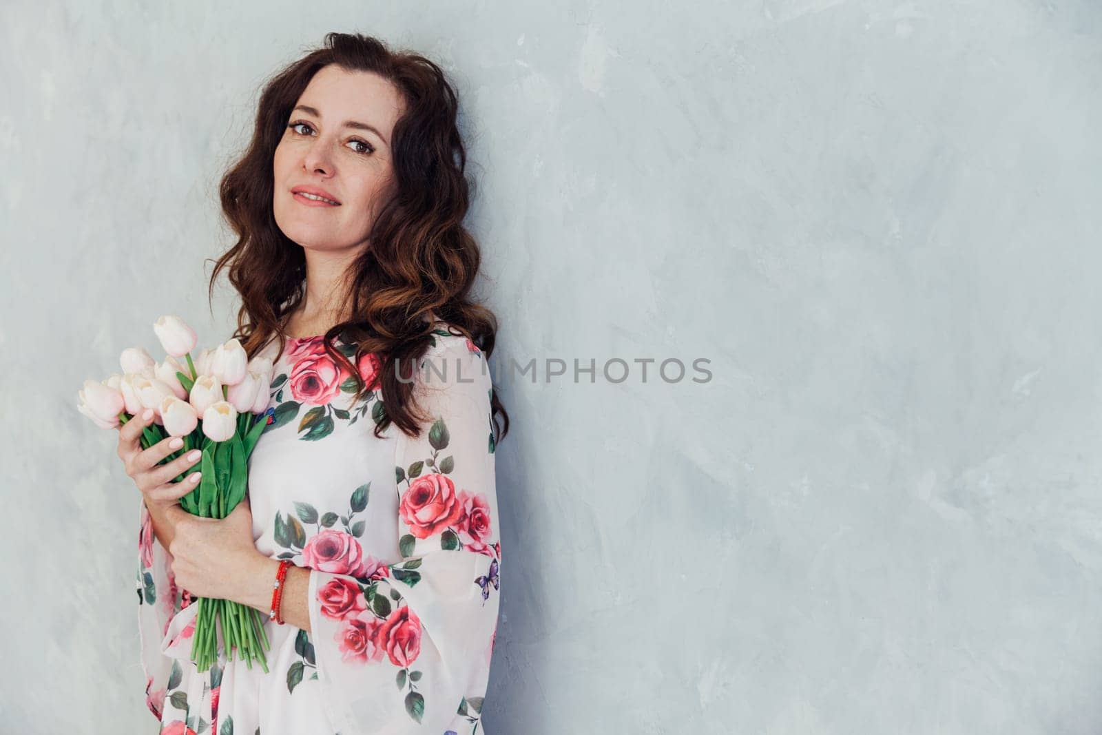 brunette woman in a colored dress with flowers in her hands stands against a gray wall holiday for a woman