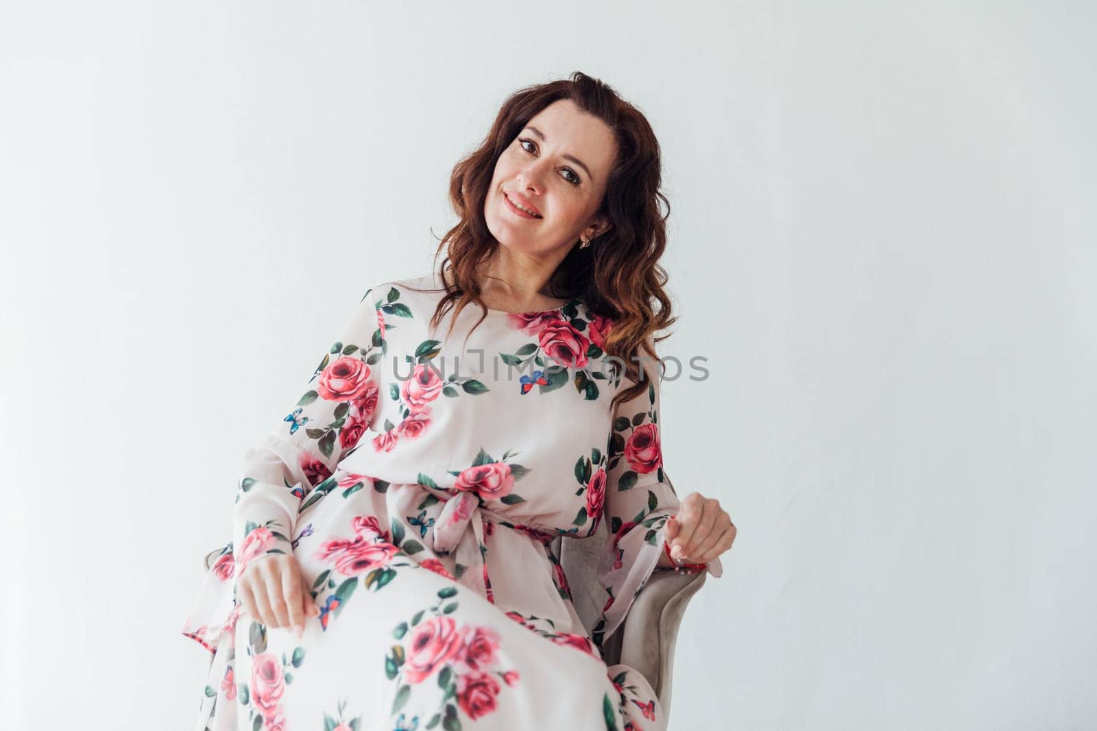 brunette woman in a floral dress sits in a chair on a light background