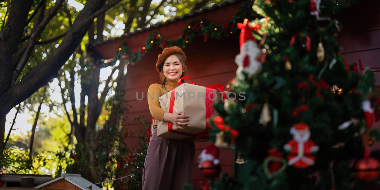 Happy woman wearing Santa hat holding of gift box. Positive emotional Santa girl. with a beautifully decorated Christmas tree serving as the background. festive Xmas concept.