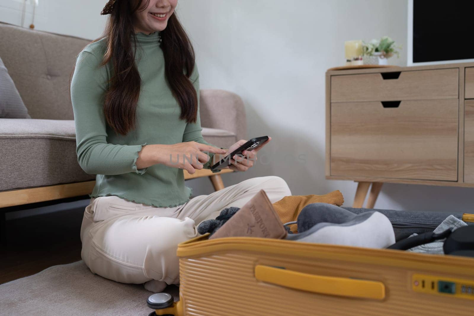 Young pretty traveler woman using smartphone to book a hotel while preparing travel suitcase.