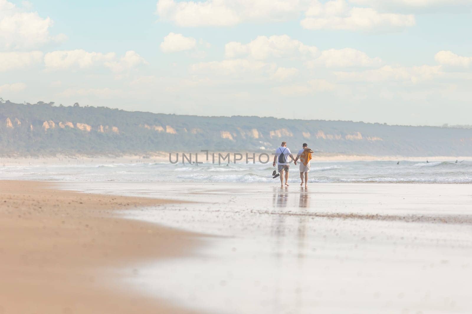 A couple of men holding his hands and walking along a beach next to the ocean - telephoto