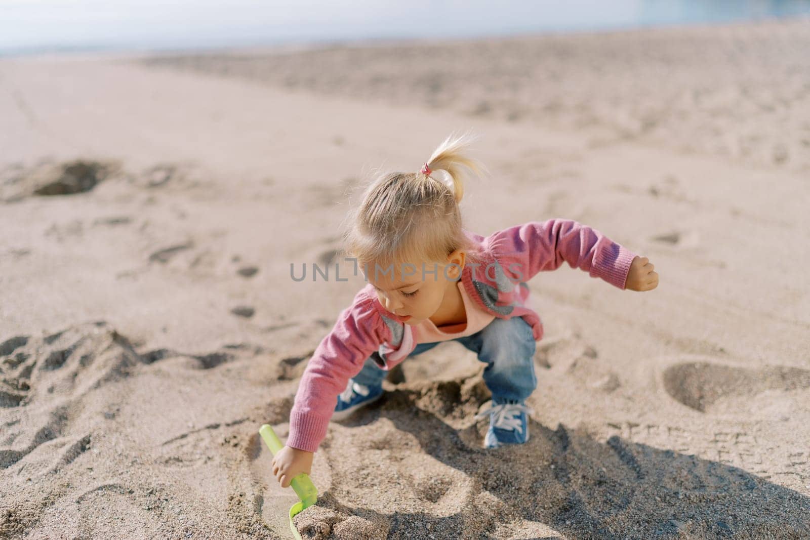 Little girl squats on the beach and digs the sand with a toy shovel with her hand out to the side. High quality photo